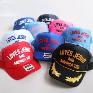 May include: A collection of trucker caps in various colours including red, black, blue, white, and pink. Each cap features the embroidered text LOVES JESUS AND AMERICA TOO in contrasting colours. Some caps include gold leaf embroidery on the brim.