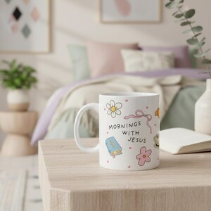 Cute Christian coffee mug styled on a bedside table in a pastel bedroom, featuring “mornings with Jesus” text, hand-drawn flowers, Bible illustration, pink bow, and tiny pink hearts coffee cup gift for women