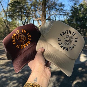 Może przedstawiać: Dwie czapki typu trucker, jedna brązowa i jedna beżowa, trzymane na tle rozmytego tła zewnętrznego. Każda czapka zawiera napis "Faith It 'Til You Make It" otaczający grafikę tygrysa. Czapki mają siatkowy tył i zakrzywiony daszek.