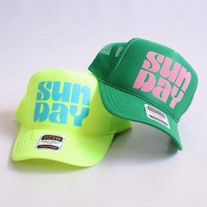 May include: Two trucker caps. One is neon yellow with the word "SUNDAY" in turquoise. The other is green with the word "SUNDAY" in pink. Both caps have a mesh back and a curved peak.