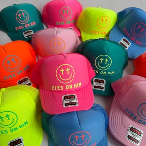 May include: A collection of vibrant trucker hats in various neon and pastel colors, each featuring a screen-printed smiley face with cross-shaped eyes and the text EYES ON HIM. Each hat includes an Otto brand label on the front foam panel.
