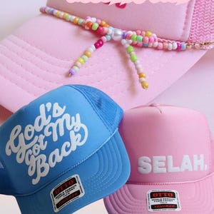Advertisement image featuring several Christian and faith-inspired products: a coffee mug with hand-drawn doodles of a Bible, flower, bow, and hearts that reads “mornings with Jesus,” a pink trucker hat with a multicolored beaded bow hat chain, a pink “Selah” hat, a light blue “God’s Got My Back” hat, and a cute notebook with hand-drawn doodles that says “mornings with Jesus.”