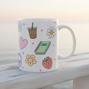 Whimsical white coffee mug with pastel hand-drawn doodles of iced coffee, hearts, journal, flower, and strawberry, styled on a white railing with beach background for cozy mornings with jesus on the beach coffee mug