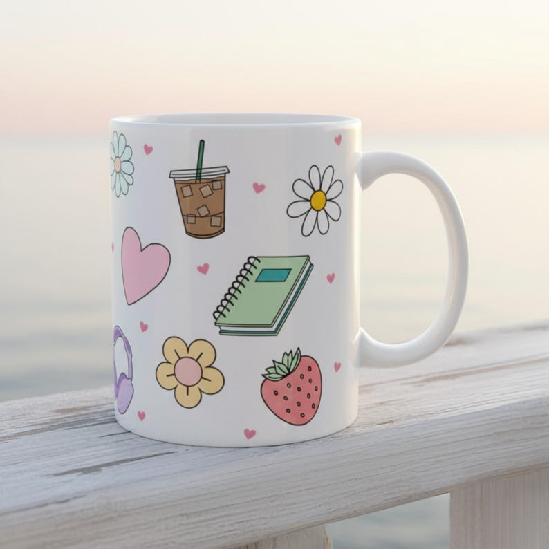 Whimsical white coffee mug with pastel hand-drawn doodles of iced coffee, hearts, journal, flower, and strawberry, styled on a white railing with beach background for cozy mornings with jesus on the beach coffee mug