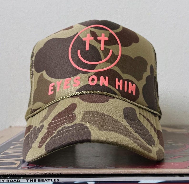 May include: A camouflage trucker hat in shades of olive green and dark brown. The front panel features a neon pink graphic of a smiley face with cross-shaped eyes and the text EYES ON HIM in neon pink lettering. A thin cord runs across the brim.
