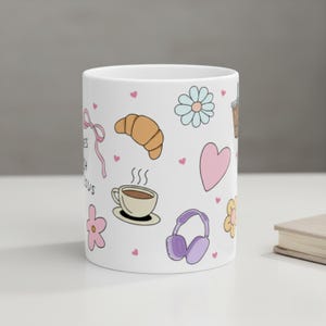 White ceramic mug on desk with cute hand drawn doodles featuring a croissant, steamy coffee cup, pink flower, purple headphones, pink flower and scattered pink mini hearts coffee cup for teen baptism gift idea
