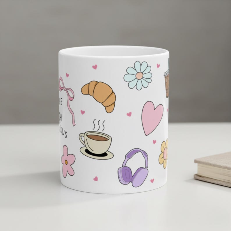 White ceramic mug on desk with cute hand drawn doodles featuring a croissant, steamy coffee cup, pink flower, purple headphones, pink flower and scattered pink mini hearts coffee cup for teen baptism gift idea