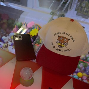 May include: A white baseball cap with a red brim. The front of the cap has a tiger graphic and the text "Jesus is my rock, He is how I roll".