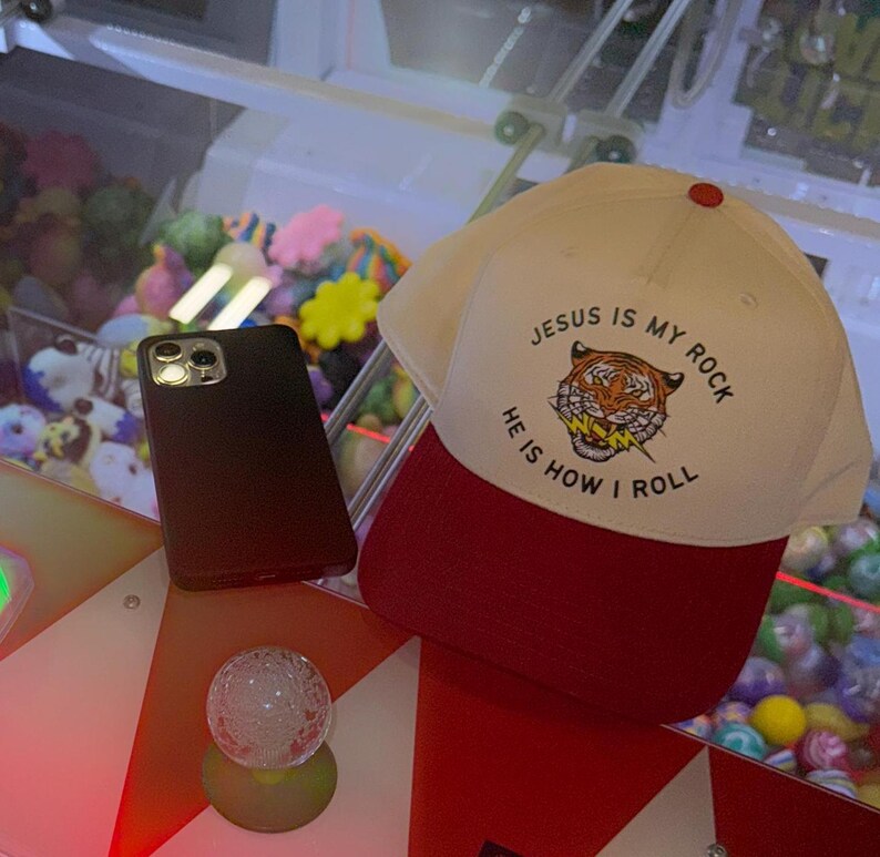 May include: A white baseball cap with a red brim. The front of the cap has a tiger graphic and the text "Jesus is my rock, He is how I roll".