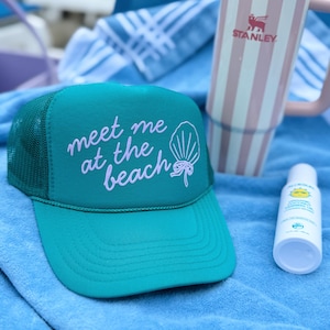 Close-up of teal Meet Me at the Beach trucker hat with seashell and bow design coastal cowgirl style summer vacation hat sitting on towel outside Beach Day Hat for Teen Girl Cute Hat for Summer