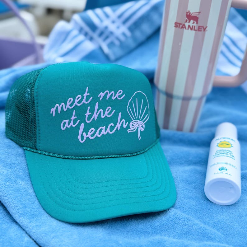 Close-up of teal Meet Me at the Beach trucker hat with seashell and bow design coastal cowgirl style summer vacation hat sitting on towel outside Beach Day Hat for Teen Girl Cute Hat for Summer