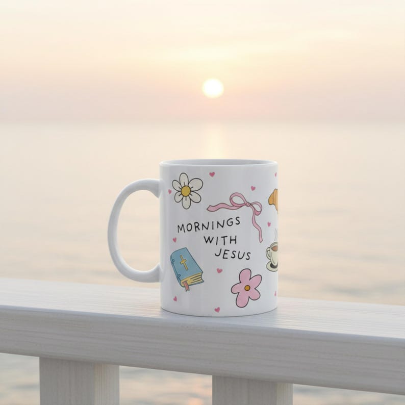 White Christian coffee mug on railing with beach background, featuring “Mornings with Jesus” text and pastel doodle Bible, flowers, pink bow, and scattered hearts.