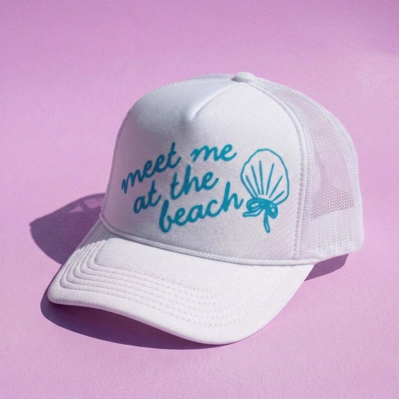 White trucker hat with teal 'meet me at the beach' script design in puff vinyl featuring a bow trucker hat with seashell