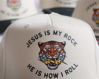 May include: A cream-colored baseball cap with a black brim. The front features a graphic of a tiger head with yellow lightning bolts in its mouth. The text "JESUS IS MY ROCK" is printed above the tiger, and "HE IS HOW I ROLL" is printed below.