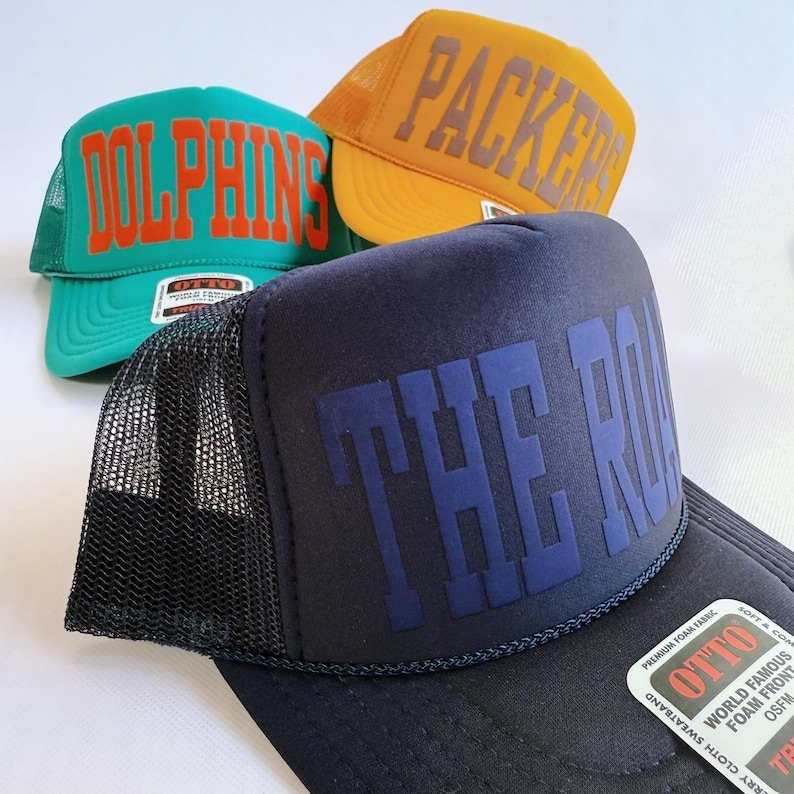 May include: Three trucker hats featuring mesh backs and foam fronts. One is teal with orange text reading DOLPHINS, one is yellow with grey text reading PACKERS, and one is black with dark blue text reading THE ROAR. Each hat has an OTTO brand label.