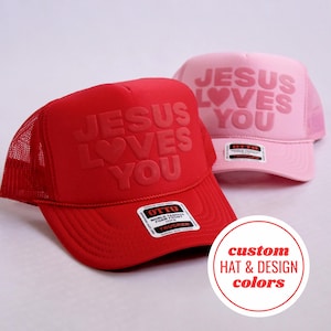 Op de afbeelding: Een felrode truckerpet en een lichtroze truckerpet, beide voorzien van de in reliëf gemaakte tekst JESUS LOVES YOU met een hartsymbool. Elke pet heeft een schuimrubberen voorkant, een mesh achterkant en een rechthoekige Otto-merksticker op de klep.
