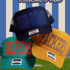 May include: Three trucker hats featuring custom text including THE ROAD, DOLPHINS, and PACKERS. The hats are navy blue, teal, and yellow with mesh backs and Otto brand labels on the front. A brown football sits on a green field with white lines.