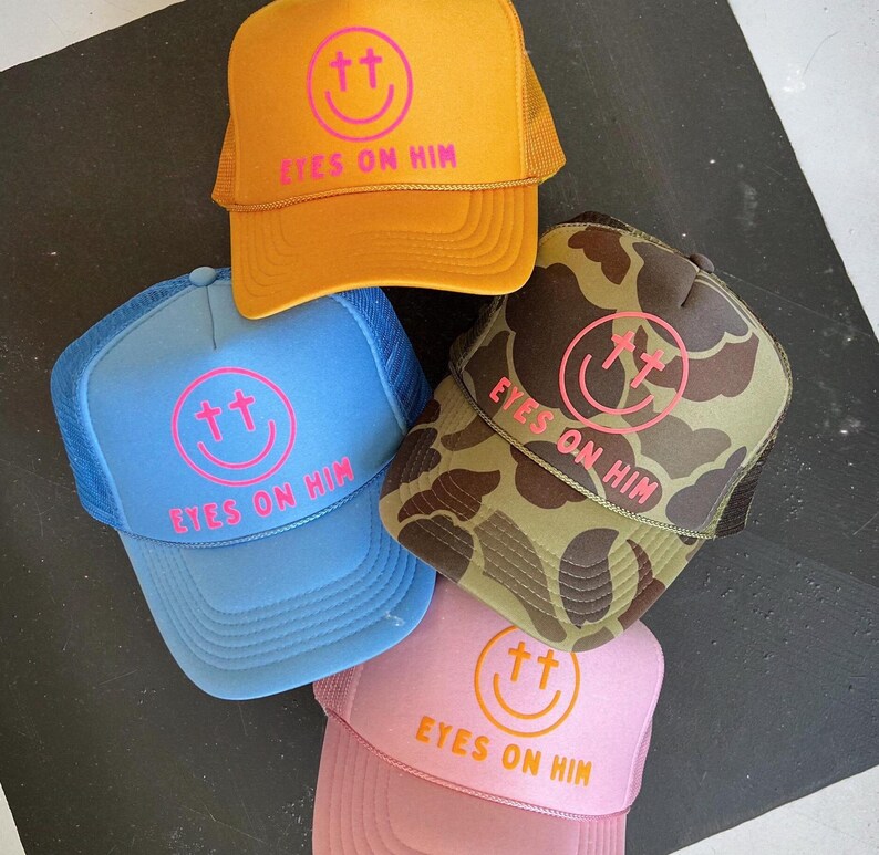 May include: Four trucker hats in yellow, light blue, camouflage, and pink. Each hat features a front graphic of a pink or orange smiley face with cross-shaped eyes and the text EYES ON HIM printed in bold capital letters below the smiley face.