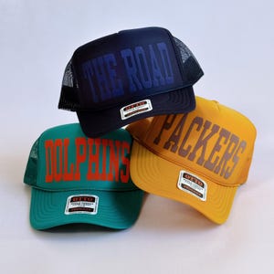 以下が含まれることがあります： ネイビー、ティール、イエローの3つのトラッカーハット。ネイビーの帽子には青いブロック体で「THE ROAD」と書かれています。ティールの帽子にはオレンジ色で「DOLPHINS」と書かれています。イエローの帽子には茶色で「PACKERS」と書かれています。