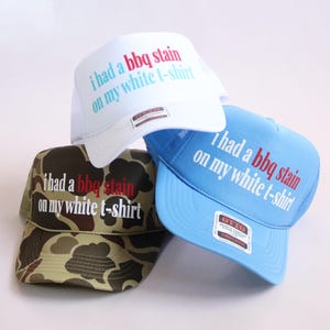 May include: Three trucker hats in white, light blue, and camouflage print. Each hat features the text "i had a bbq stain on my white t-shirt" printed on the front panel in red and white or light blue lettering. The hats have mesh back panels.