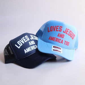 May include: Two foam front trucker hats with mesh backs. The navy blue hat features white text reading LOVES JESUS AND AMERICA. The light blue hat features red text reading LOVES JESUS AND AMERICA TOO, with an Otto brand sticker on the brim.