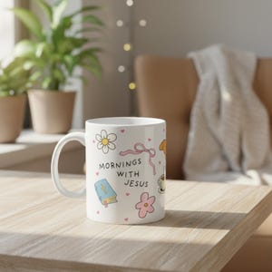Faith-inspired coffee mug with “mornings with Jesus” design, hand-drawn flowers, Bible, pink bow, and hearts, styled on a bedside table in a soft neutral bedroom.
