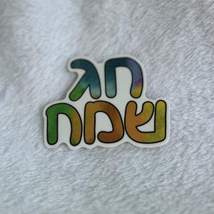 May include: A colorful sticker with the Hebrew words "Chag Sameach" in a rainbow tie-dye design. The sticker has a white border and is set against a soft, white, textured background.