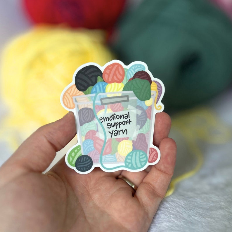 May include: A sticker of a clear container filled with colorful yarn balls. The container has a label that reads "Emotional Support Yarn".