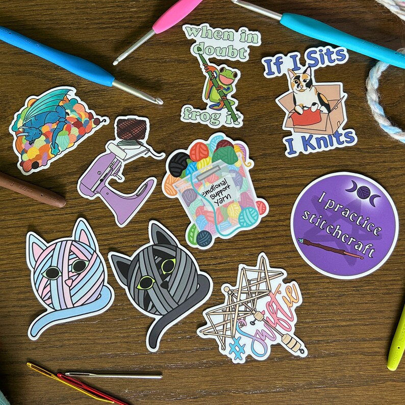 May include: A collection of colorful stickers featuring various knitting and crocheting themes. The stickers include a cat wrapped in yarn, a frog, a yarn bowl, a knitting needle, and a cat sitting in a box with the text "If I sits I knits".