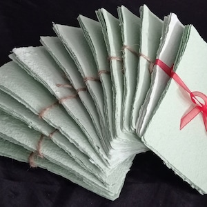 May include: A stack of handmade, light green paper sheets with rough edges, tied together with twine and decorated with a red ribbon bow.