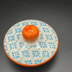 May include: A ceramic lid with a floral pattern. The lid is white with blue flower designs and an orange rim. A small orange knob is centered on top. The lid is likely for a teapot or similar container.
