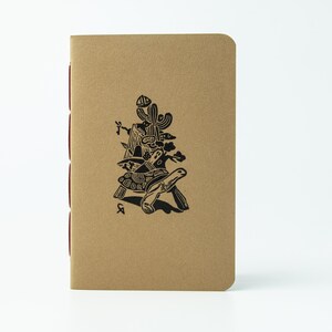 Puede incluir: Un cuaderno beige con una ilustración negra de una tortuga que lleva una escena desértica, incluyendo un cactus y una montaña. El cuaderno tiene un lomo rojo y un diseño sencillo y artístico.