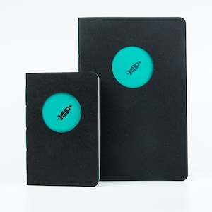 Può includere: Due quaderni neri di diverse dimensioni. Ogni quaderno ha un ritaglio circolare che rivela un interno turchese con un'icona di razzo nero. I quaderni sono su uno sfondo bianco.