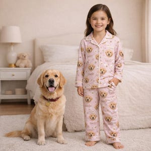 May include: A young girl wearing pink pajamas with a repeating pattern of golden retriever faces and the name "Daisy". A golden retriever dog sits beside her. The pajamas have white trim and the word "Daisy" is also printed on the pants.