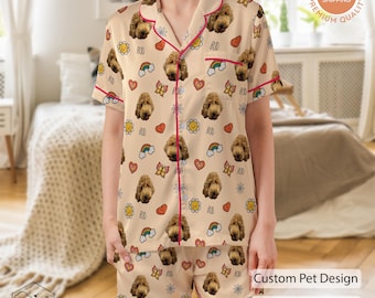 May include: Beige pyjama set featuring a repeating pattern of a dog's face, rainbows, hearts, and flowers. The set includes a short-sleeved button-up shirt and matching shorts, trimmed with red piping. The text "Custom Pet Design" and "Premium Quality" is visible.