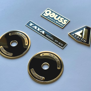 Könnte beinhalten: Eine Auswahl an metallischen Abzeichen und Emblemen. Zwei kreisförmige schwarz-goldene Embleme mit den Worten "BRILLIANCE" und "INCREASE" sind vorhanden. Rechteckige Abzeichen sind "gauss", "TAYA MODEL DP-S10" und ein dreieckiges Emblem "AVALON".