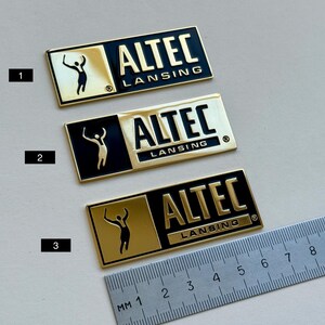 K&ouml;nnte beinhalten: Drei rechteckige gold-schwarze ALTEC LANSING-Abzeichen. Jedes Abzeichen zeigt den Markennamen und eine stilisierte Figur. Ein Lineal mit Millimeter-Markierungen befindet sich im Bild.