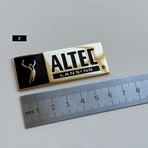 K&ouml;nnte beinhalten: Ein rechteckiges, goldfarbenes Metallabzeichen mit dem Text "ALTEC LANSING" in Schwarz. Das Abzeichen hat einen schwarzen Bereich mit einer goldenen Figur. Ein Lineal liegt neben dem Abzeichen. Das Abzeichen ist etwa 7,6 cm lang.