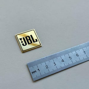 Peut inclure: Un badge carr&eacute; avec le logo JBL, bordure dor&eacute;e et centre cr&egrave;me. Le texte JBL noir est &agrave; c&ocirc;t&eacute; d'une r&egrave;gle argent&eacute;e avec des graduations en millim&egrave;tres. Le badge mesure environ 2,5 cm de c&ocirc;t&eacute;.