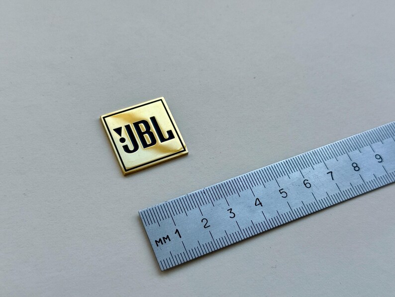 Peut inclure: Un badge carr&eacute; avec le logo JBL, bordure dor&eacute;e et centre cr&egrave;me. Le texte JBL noir est &agrave; c&ocirc;t&eacute; d'une r&egrave;gle argent&eacute;e avec des graduations en millim&egrave;tres. Le badge mesure environ 2,5 cm de c&ocirc;t&eacute;.