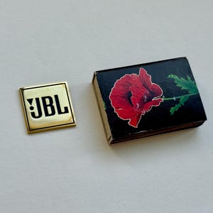 Peut inclure: Une &eacute;pingle carr&eacute;e dor&eacute;e avec les lettres "JBL" en noir. &Agrave; c&ocirc;t&eacute;, une bo&icirc;te d'allumettes noire avec une illustration de fleur de pavot rouge. La bo&icirc;te a un contour blanc autour de la fleur et des feuilles vertes.
