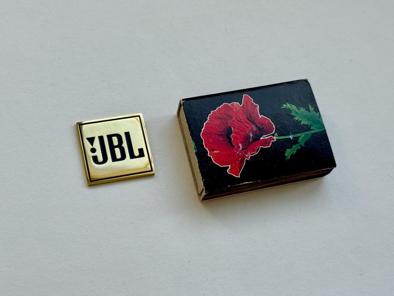 Peut inclure: Une &eacute;pingle carr&eacute;e dor&eacute;e avec les lettres "JBL" en noir. &Agrave; c&ocirc;t&eacute;, une bo&icirc;te d'allumettes noire avec une illustration de fleur de pavot rouge. La bo&icirc;te a un contour blanc autour de la fleur et des feuilles vertes.