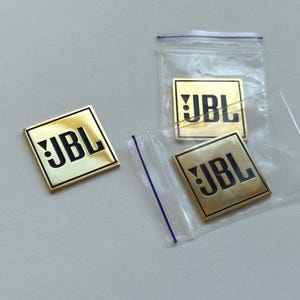 Può includere: Diversi distintivi dorati con il logo JBL. I distintivi quadrati hanno un logo JBL nero. Due distintivi sono in sacchetti di plastica trasparente. I distintivi sono su una superficie grigio chiaro.