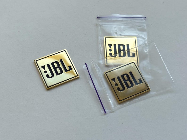 Peut inclure: Plusieurs badges dor&eacute;s avec le logo JBL. Les badges carr&eacute;s ont un logo JBL noir. Deux badges sont dans des sacs en plastique transparent. Les badges sont sur une surface gris clair.