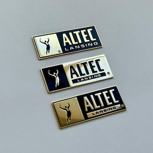 K&ouml;nnte beinhalten: Drei rechteckige Metallabzeichen mit dem Text "ALTEC LANSING" in wei&szlig;er Schrift. Jedes Abzeichen hat einen goldfarbenen Rahmen und einen schwarzen Hintergrund. Eine Silhouette einer Person mit erhobenen Armen befindet sich auf der linken Seite.