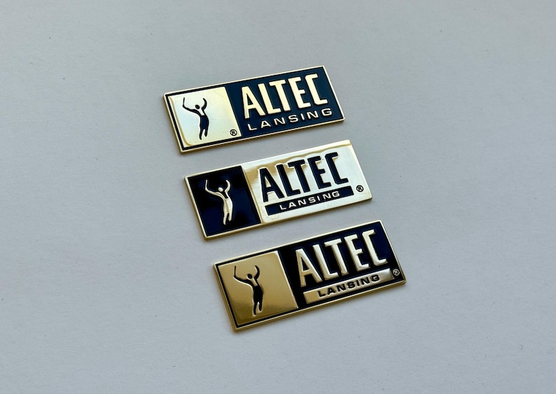 K&ouml;nnte beinhalten: Drei rechteckige Metallabzeichen mit dem Text "ALTEC LANSING" in wei&szlig;er Schrift. Jedes Abzeichen hat einen goldfarbenen Rahmen und einen schwarzen Hintergrund. Eine Silhouette einer Person mit erhobenen Armen befindet sich auf der linken Seite.