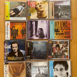 Op de afbeelding: Een verzameling van twaalf muziek-cd's in plastic doosjes, gerangschikt in een raster. De hoezen tonen diverse artiesten waaronder The Wannadies, Gangway, Roger Morris, Primal Scream, Huey Lewis, Bruce Springsteen, King Crimson, Ron Wood, Rod Stewart en Sting, velen met Japanse obi-strips.