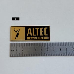 K&ouml;nnte beinhalten: Ein rechteckiges gold-schwarzes ALTEC LANSING-Abzeichen mit einer stilisierten Figur und Text. Das Abzeichen ist etwa 7,6 cm lang, mit einem Lineal zur Ma&szlig;stabsbestimmung. Der Text "ALTEC LANSING" ist deutlich sichtbar.