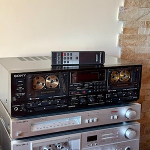 Puede incluir: Una pila de equipos de audio vintage, que incluye una pletina de casete Sony negra con un mando a distancia y dos componentes Telefunken plateados. La pletina Sony presenta dos ranuras para cintas y el texto "AUTO REVERSE".