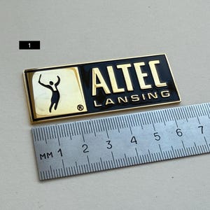 K&ouml;nnte beinhalten: Ein rechteckiges, gold-schwarzes ALTEC LANSING-Emblem. Das Emblem zeigt den Markennamen in goldfarbenen Buchstaben und eine stilisierte Figur. Ein Lineal liegt neben dem Emblem und zeigt Messungen in Millimetern.